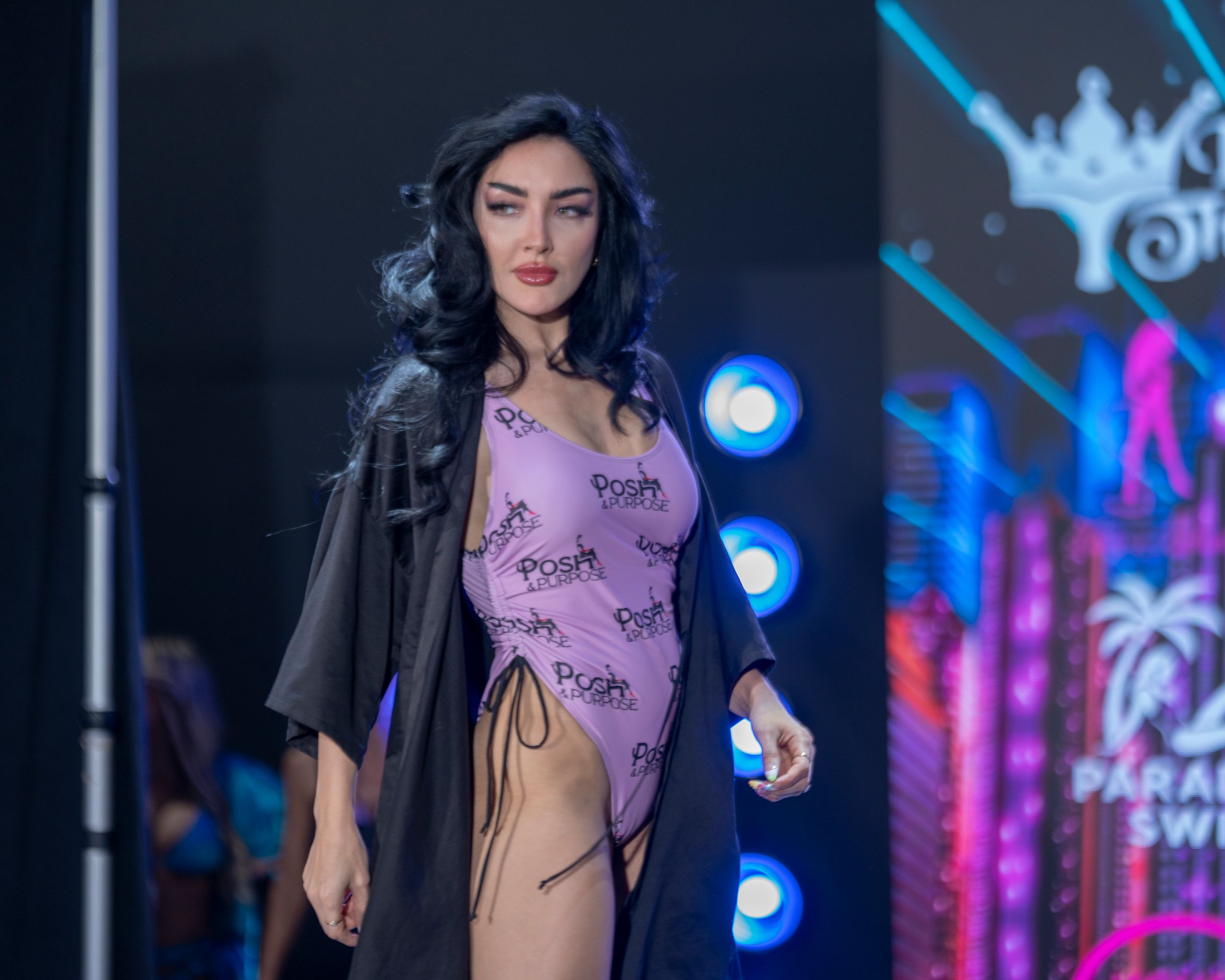 Fashion model walks runway in colorful evening dress at fashion show with vibrant neon lighting and palm tree decor.
