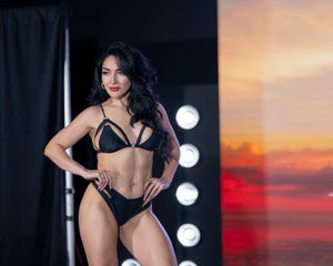 Model wearing black bikini poses confidently on stage with sunset backdrop.
