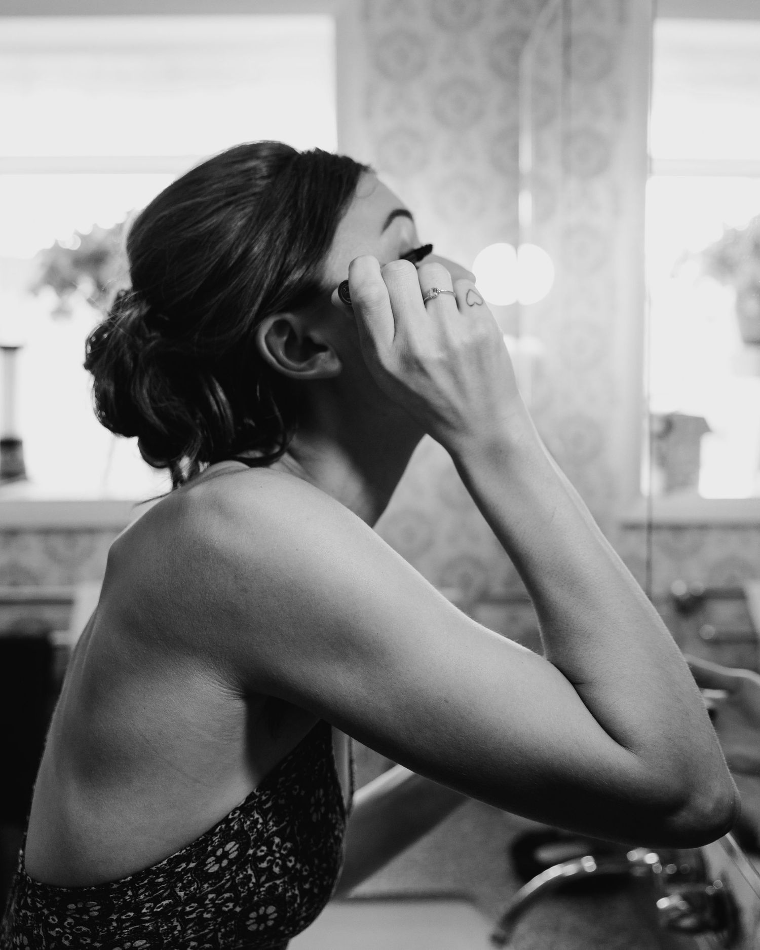 Black and white photo of a woman applying eye makeup while looking in a bathroom mirror.