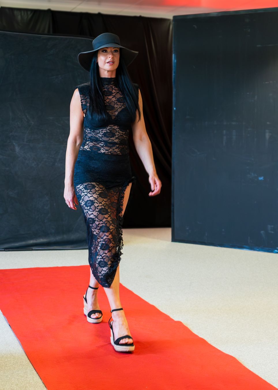 A person wearing a black lace dress and hat walks on a red carpet runway against a dark backdrop.