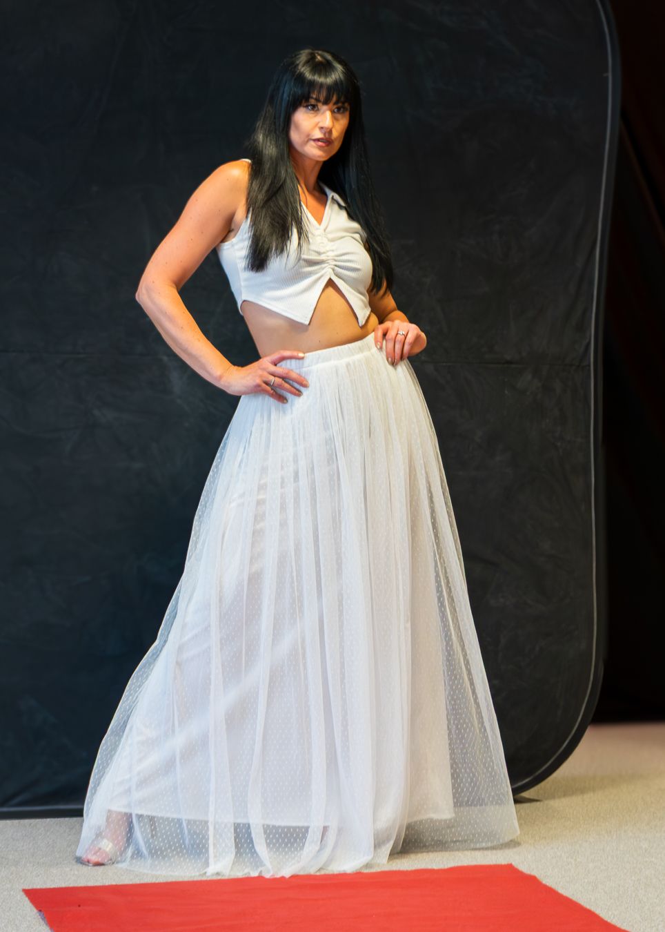 A person in a flowing white dress poses on a red carpet against a dark backdrop during a fashion photoshoot.