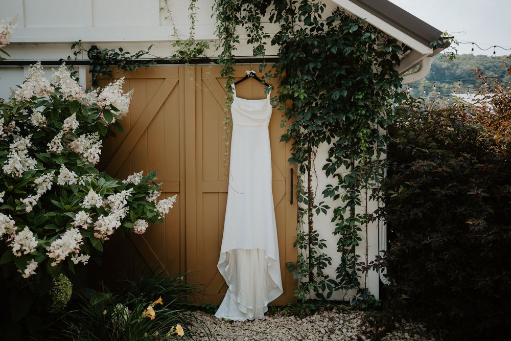 White wedding dress hanging elegantly against a rustic wooden wall with flowering vines.