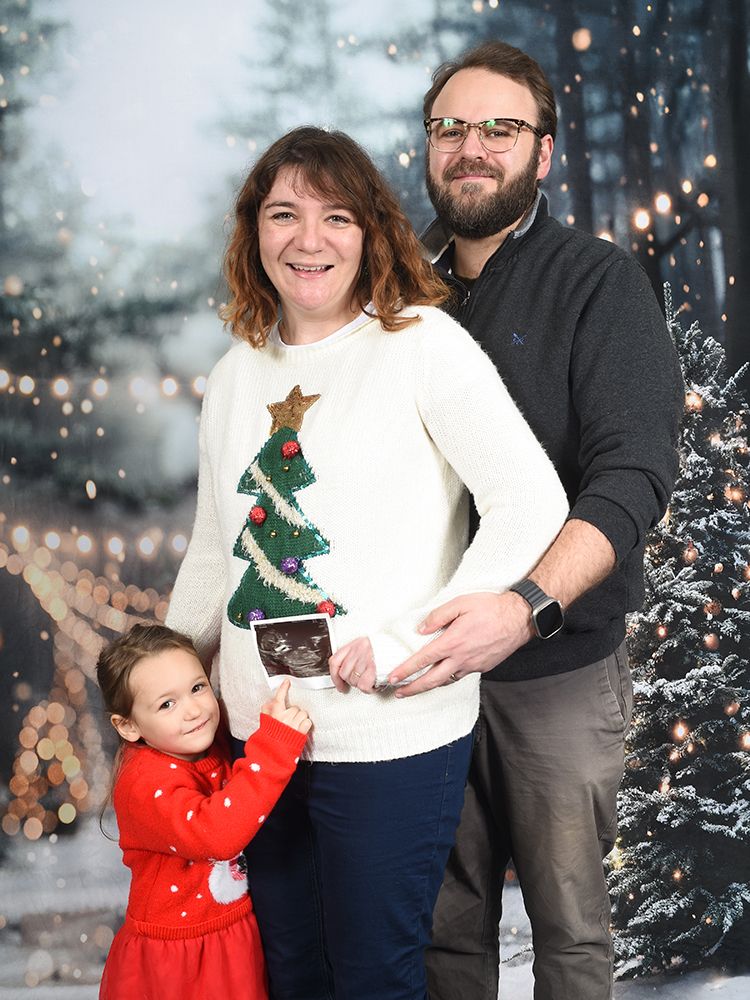 Women wearing festive jumper holds baby scan photo in family photograph on a wooded Christmas themed photoshoot at Abigail's Studio