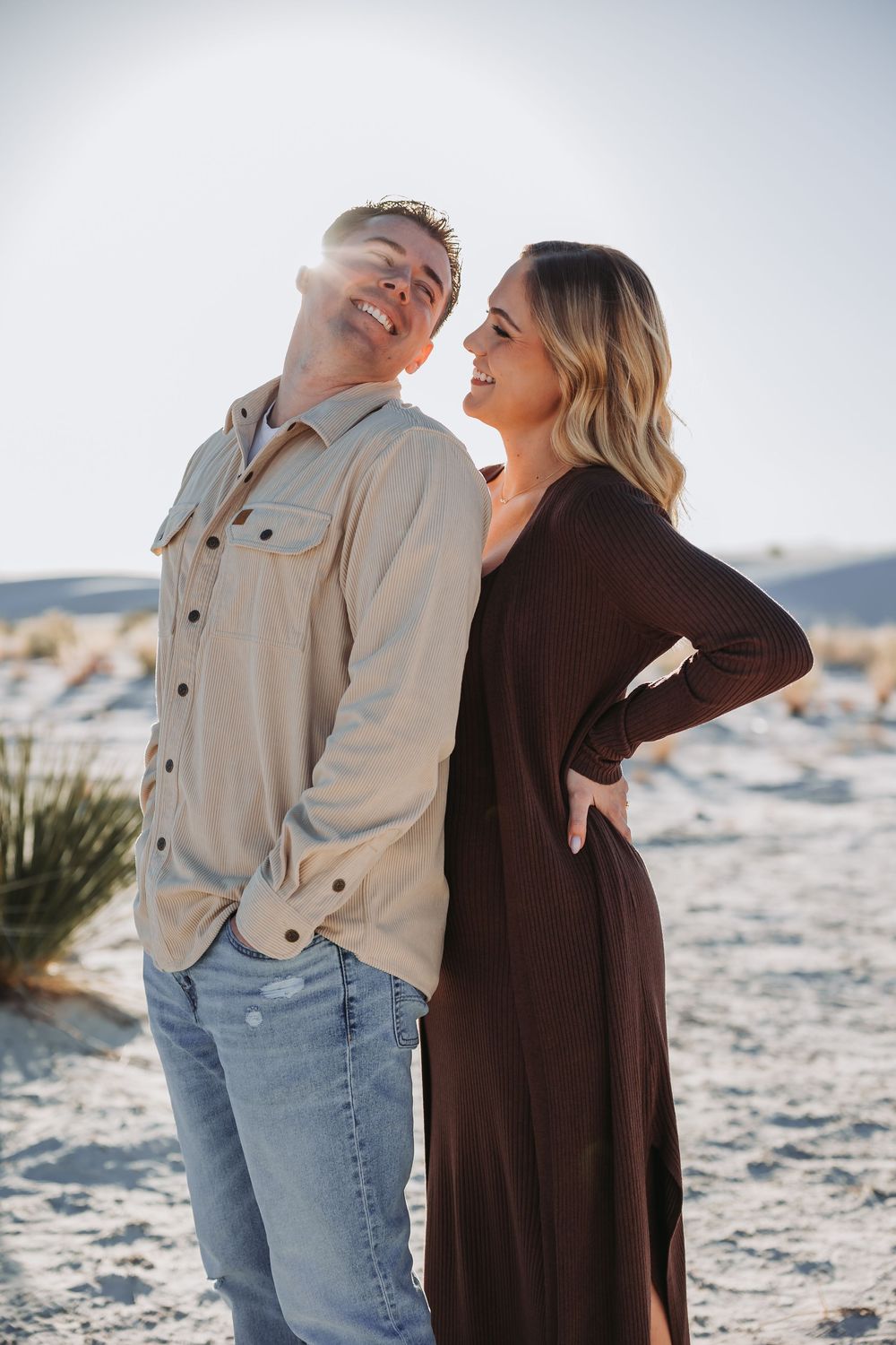 Couple in casual attire sharing playful moments together on a sunny beach with light sand and natural lighting.