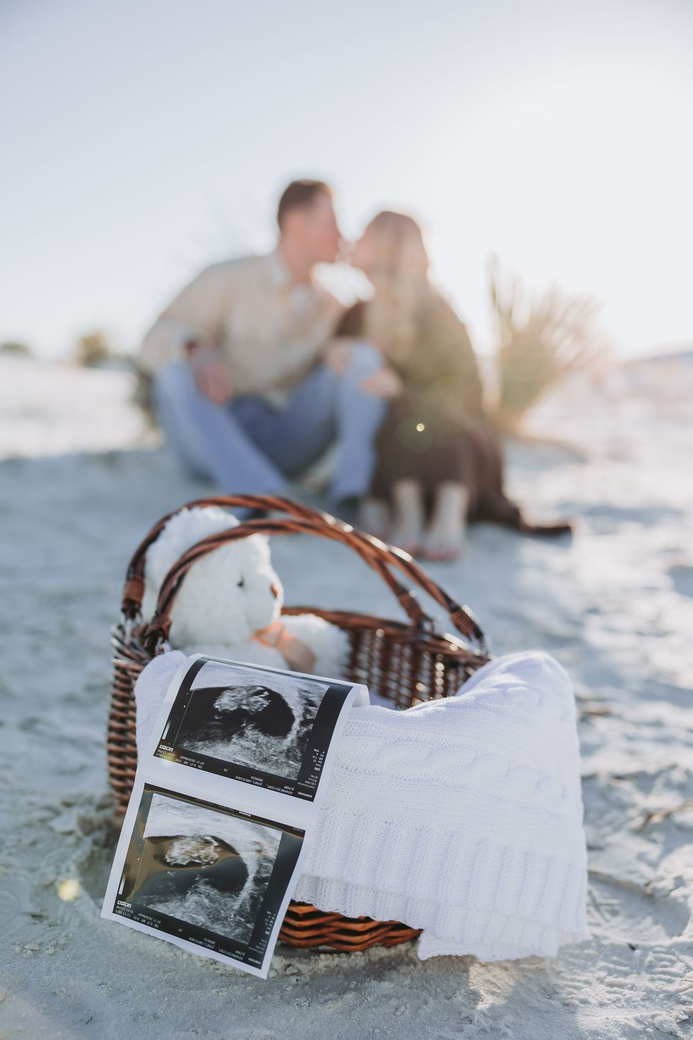 A wicker basket with ultrasound photos and baby items sits on sandy beach while couple relaxes in background.