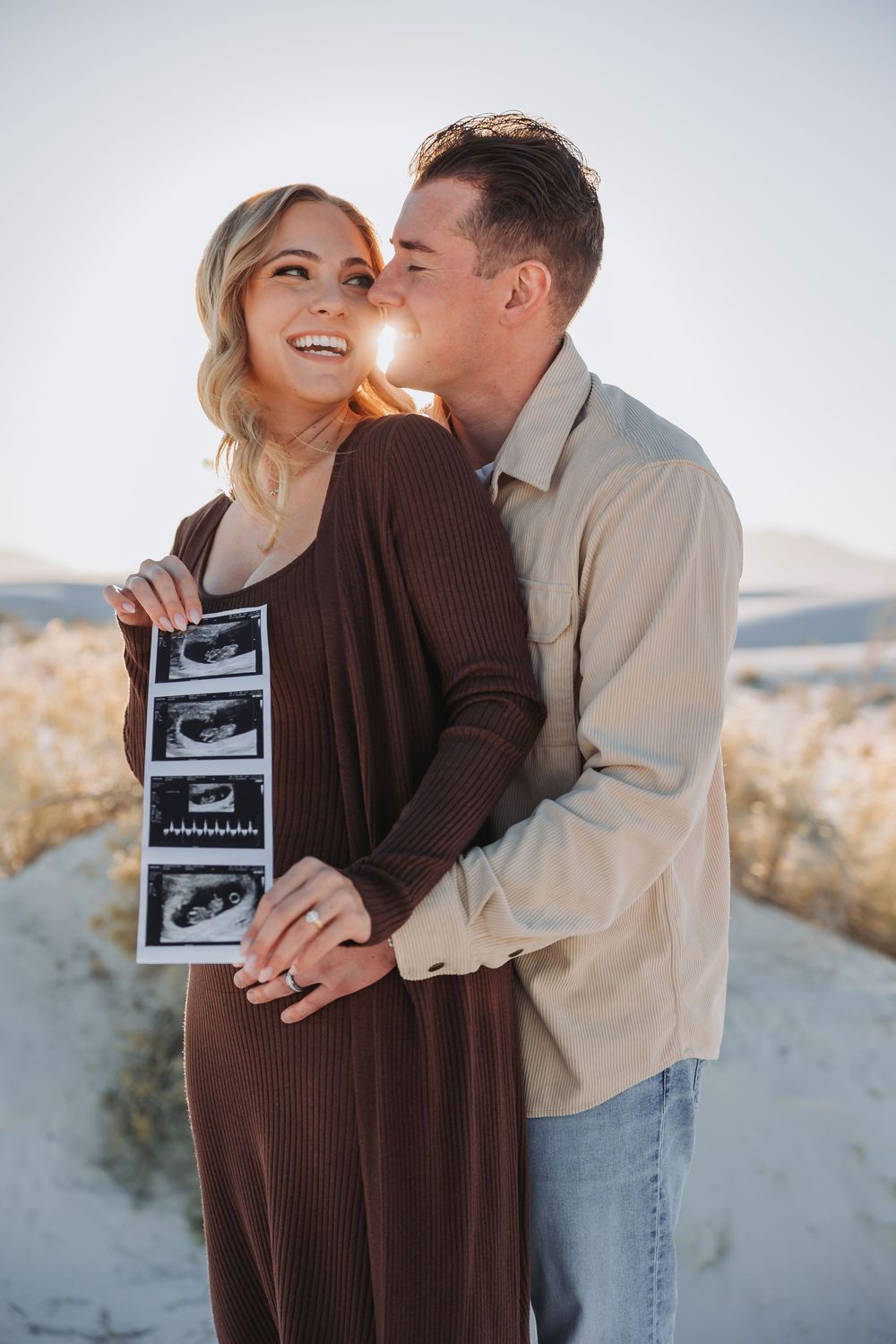 A couple shares a tender moment on a beach while holding ultrasound photos to announce their pregnancy.