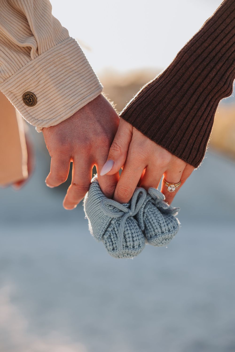Two hands holding small gray knitted baby booties while wearing rings and different sleeve styles.