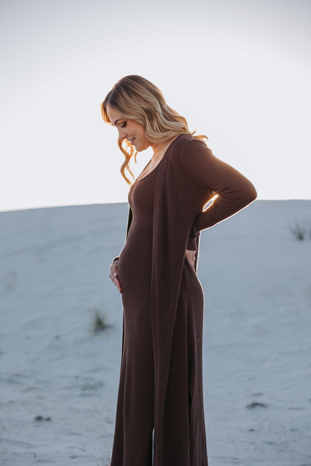 A person in a long brown dress stands against a light gray background in a contemplative pose.