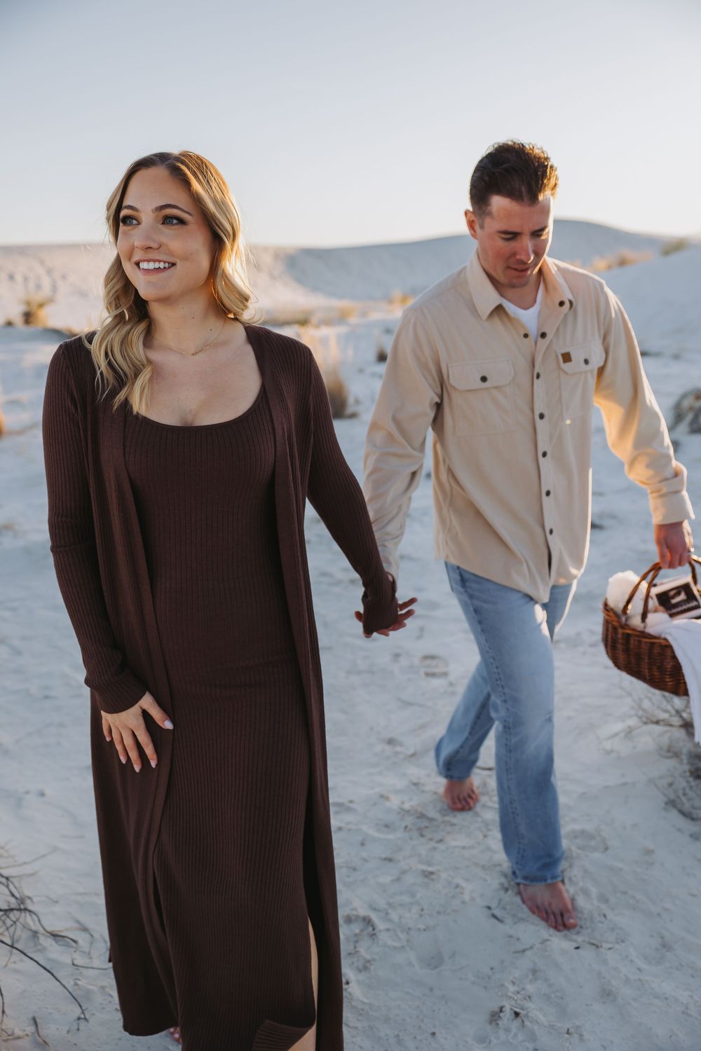 A couple walks playfully along a sandy beach at sunset wearing casual brown and beige outfits.