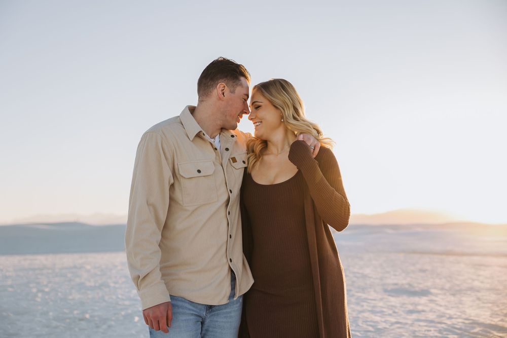 A couple embraces on the beach during golden hour with ocean waves and sunset in the background.