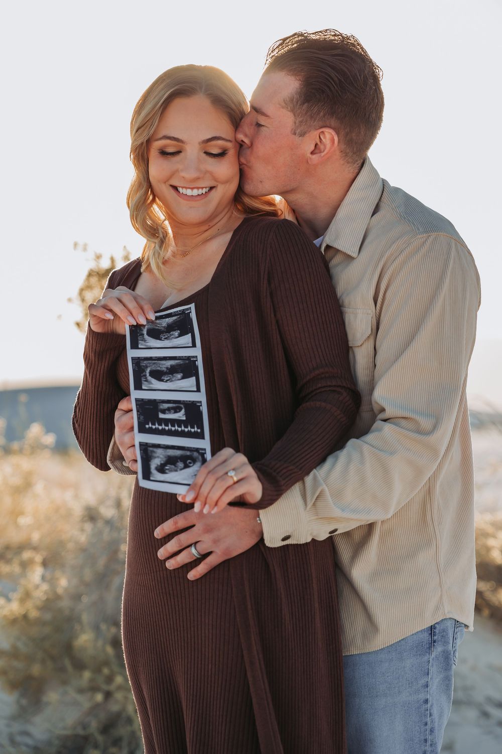 Couple sharing tender moment while holding ultrasound photos during maternity photoshoot at sunset.