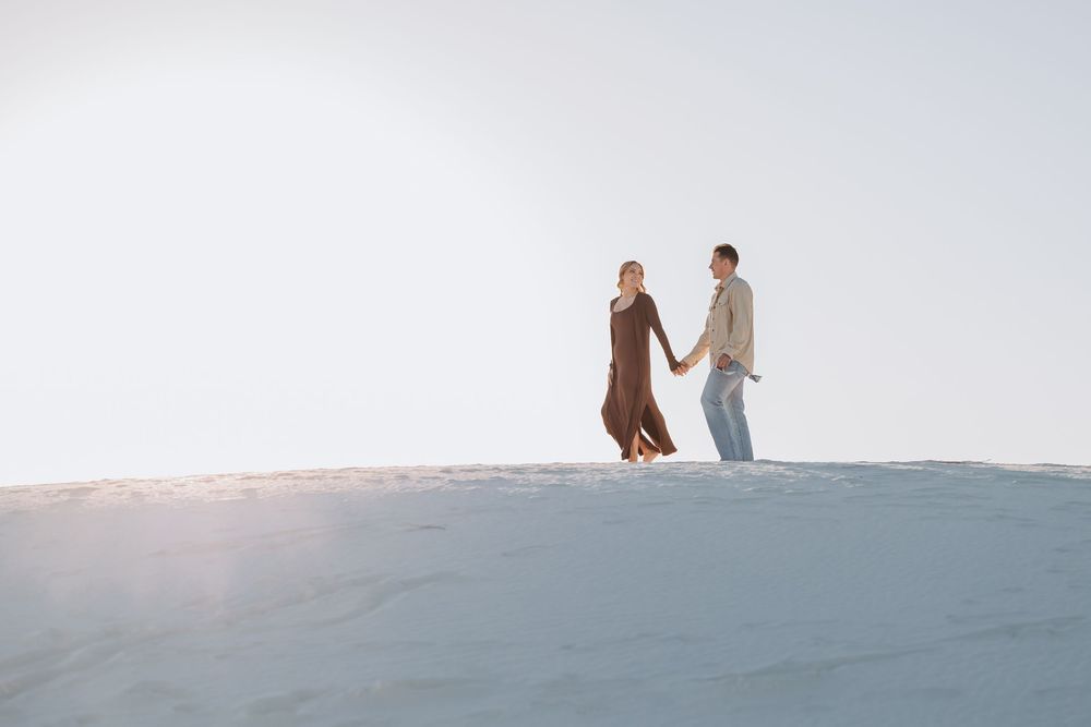 Two people walk along a snowy hill against a bright winter sky, creating silhouettes in the distance.