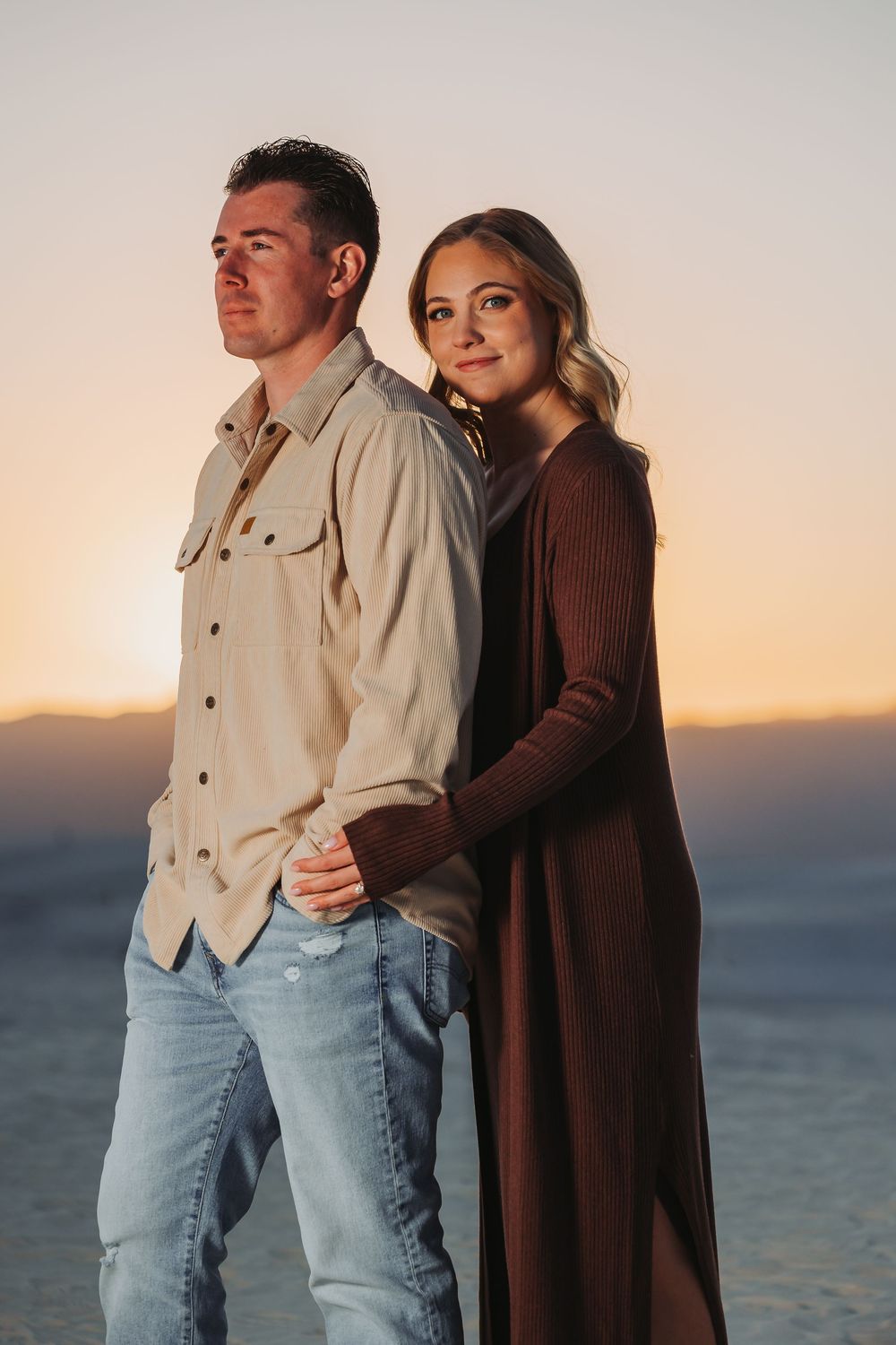 A couple in casual attire embraces while standing against an ocean backdrop during golden hour sunset.
