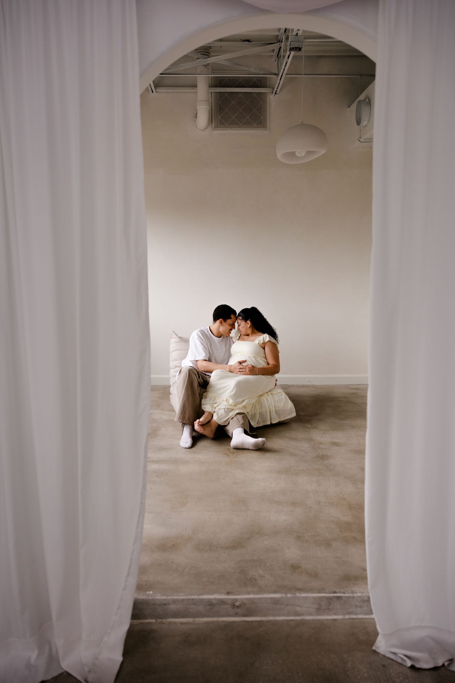 A couple sits intimately together on the floor, viewed through a curtained archway in a minimalist white space.