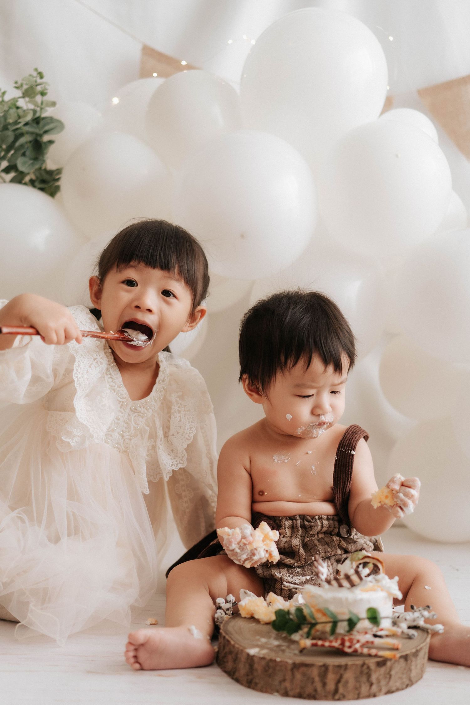 Two children enjoying cake and treats with white balloons and rustic decor at a special celebration.