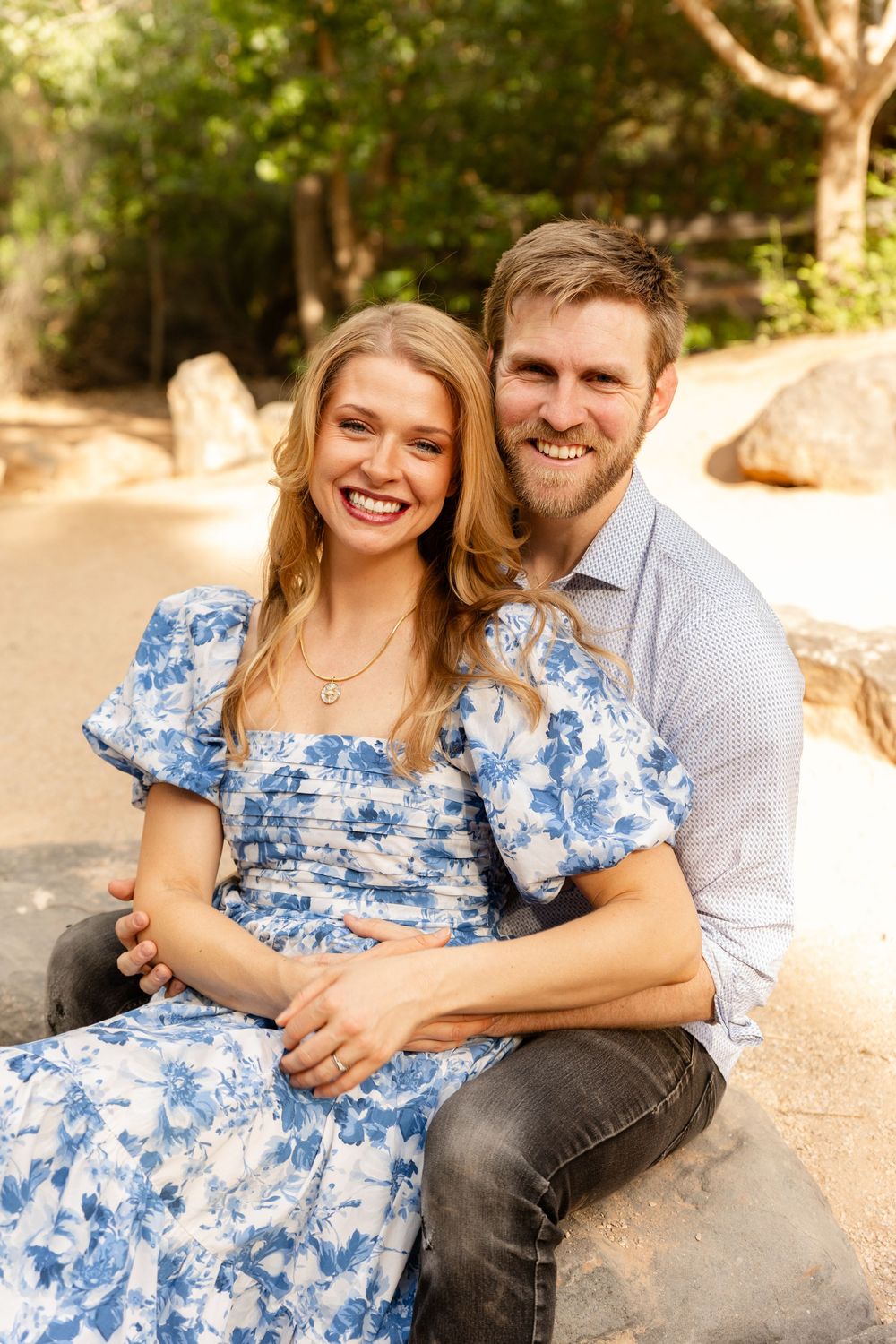 a couple in church dress smile at the camera while sitting on a rock