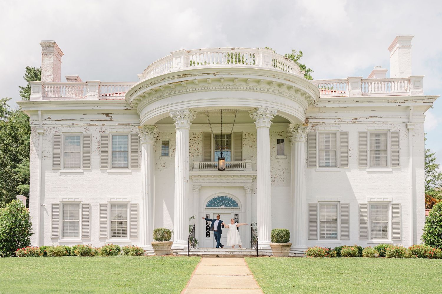 A grand white antebellum mansion with curved columned portico entrance and symmetrical windows stands majestically on green lawn.