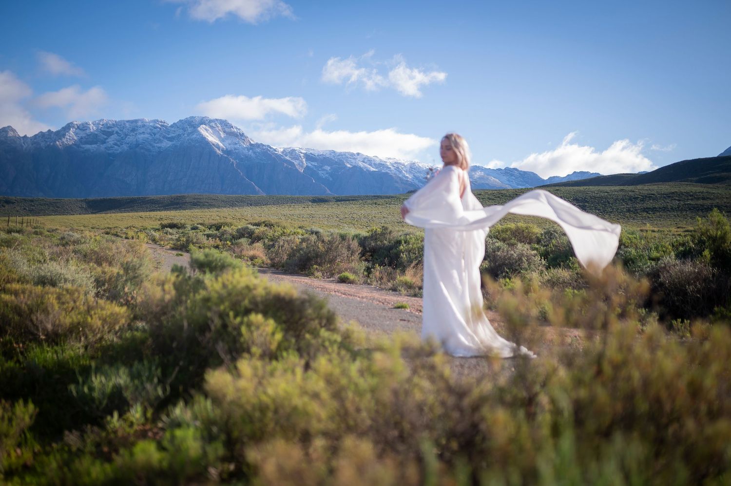 A person in white stands in a mountain sagebrush landscape under blue skies and distant snowy peaks.