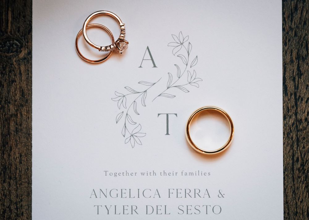 Two gold wedding rings rest on elegant white wedding invitation stationery with floral designs and calligraphy text.