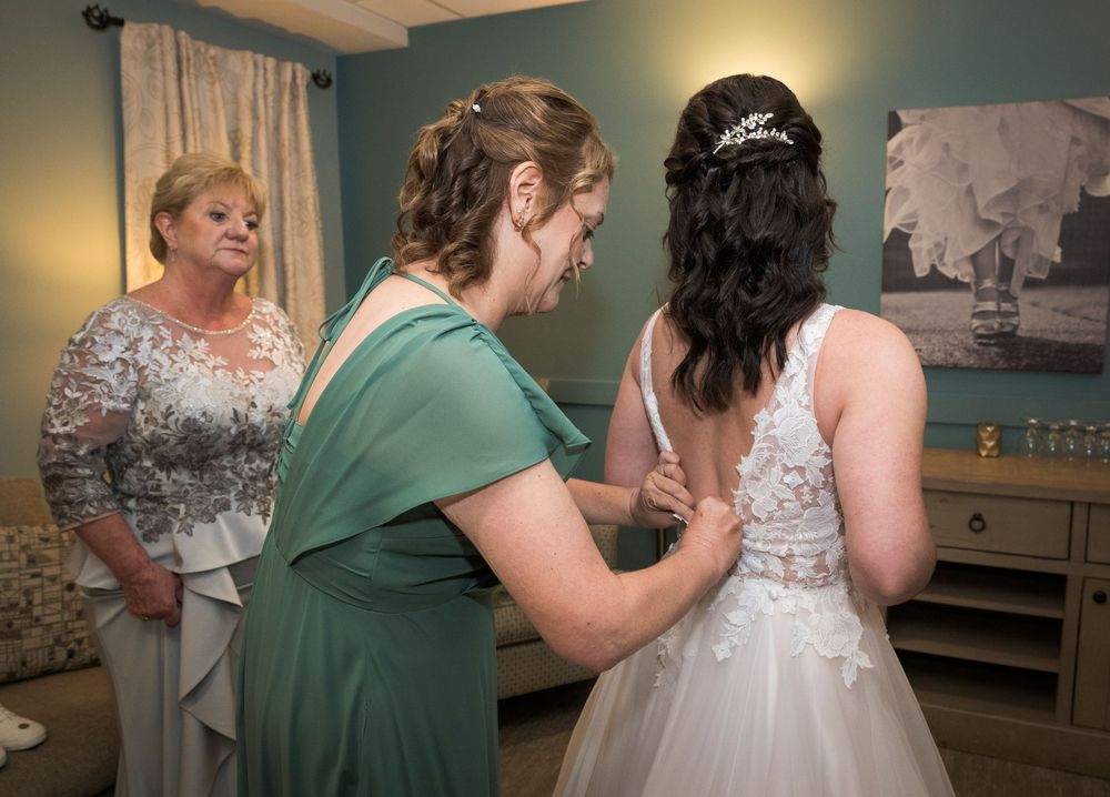 Bridesmaids in teal dresses help adjust a wedding gown in a preparation room.