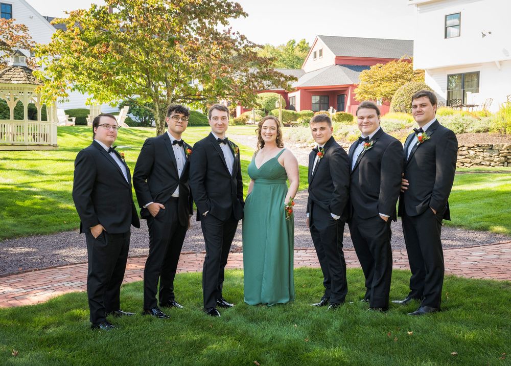Wedding party in formal attire poses outdoors with groomsmen in black tuxedos and bridesmaids in sage green dresses.