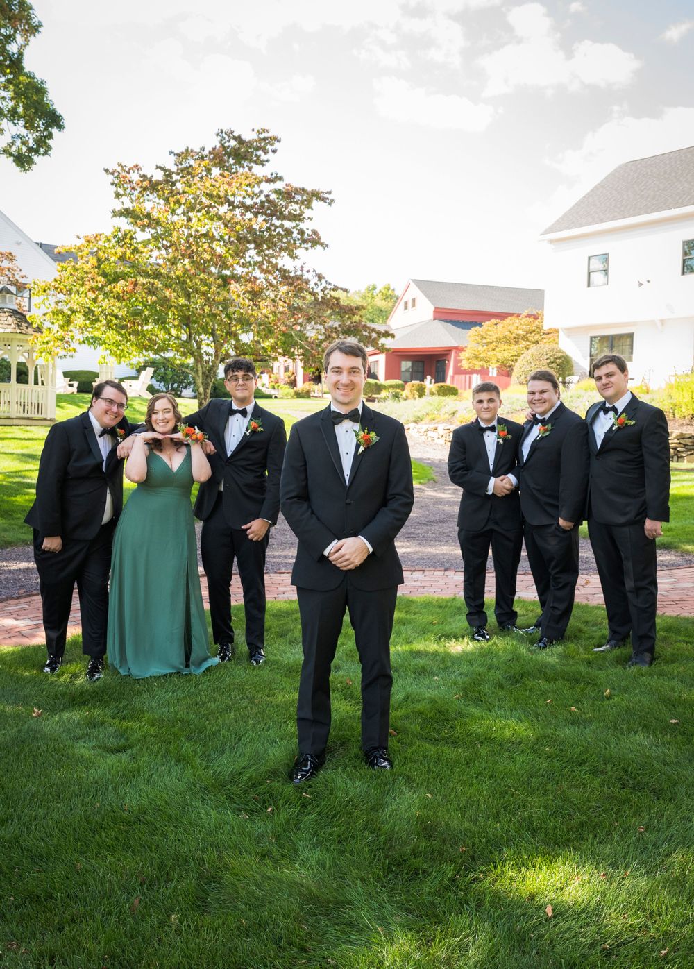 Wedding party in black suits and green dresses pose on sunny lawn for group photos.