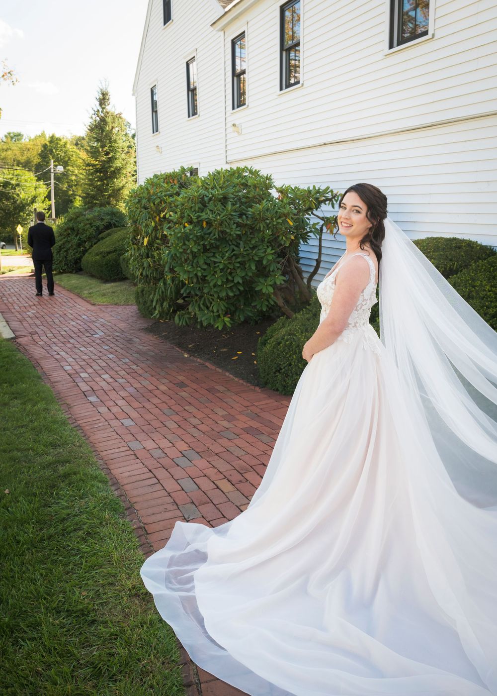 Flowing white wedding dress train cascades along a brick pathway beside a white house.