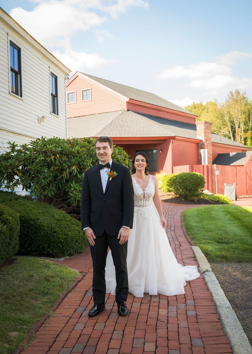 Wedding couple poses on brick pathway outside white colonial building with red barn in background.