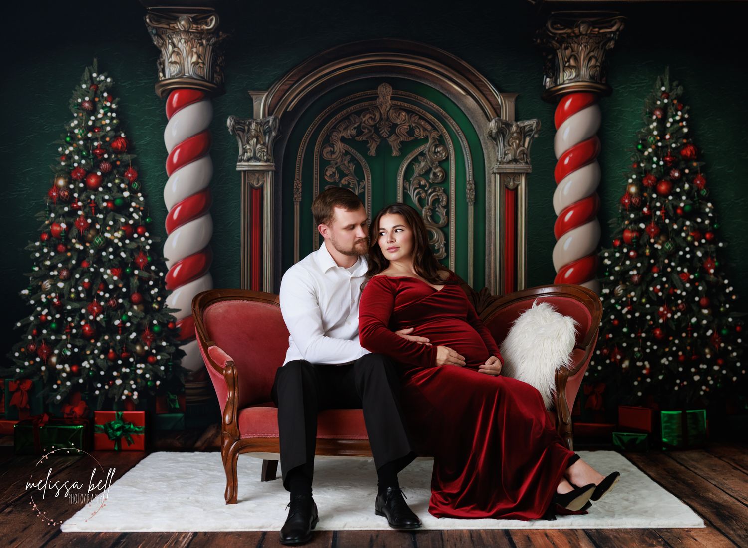 Christmas photoshoot featuring couple in white and red outfits posing in front of decorated holiday backdrop with trees.
