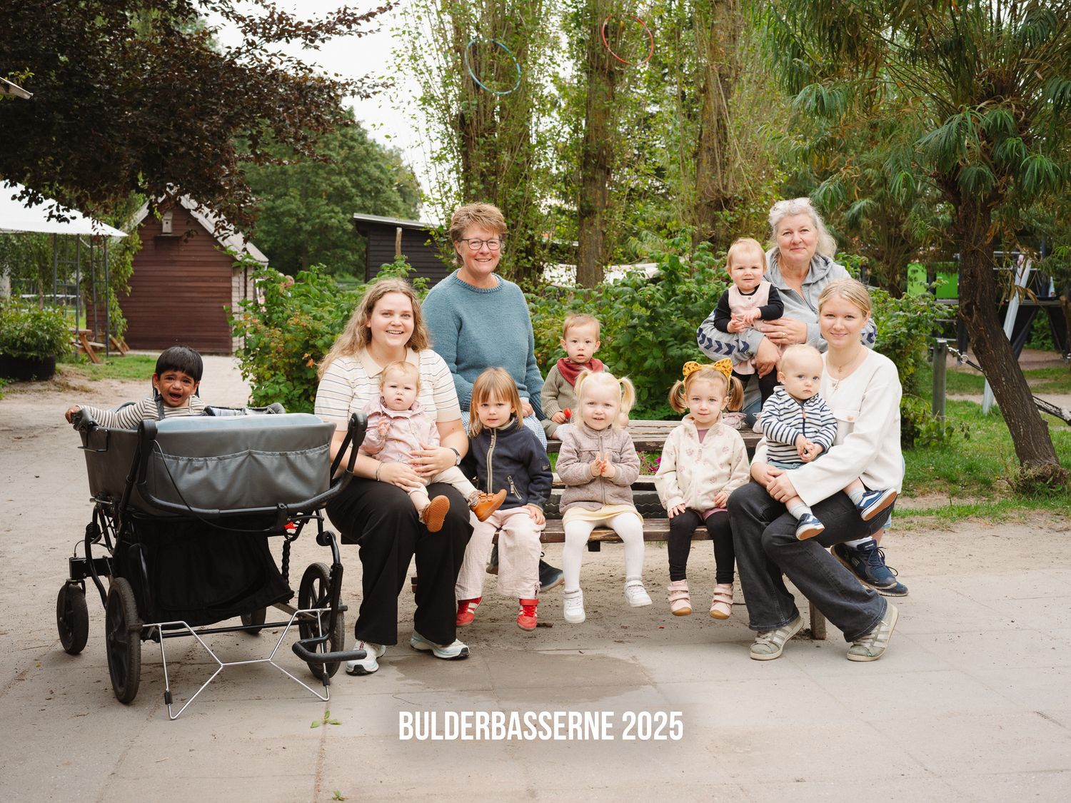 A group of people sitting together outdoors on a park bench with strollers and trees in the background.