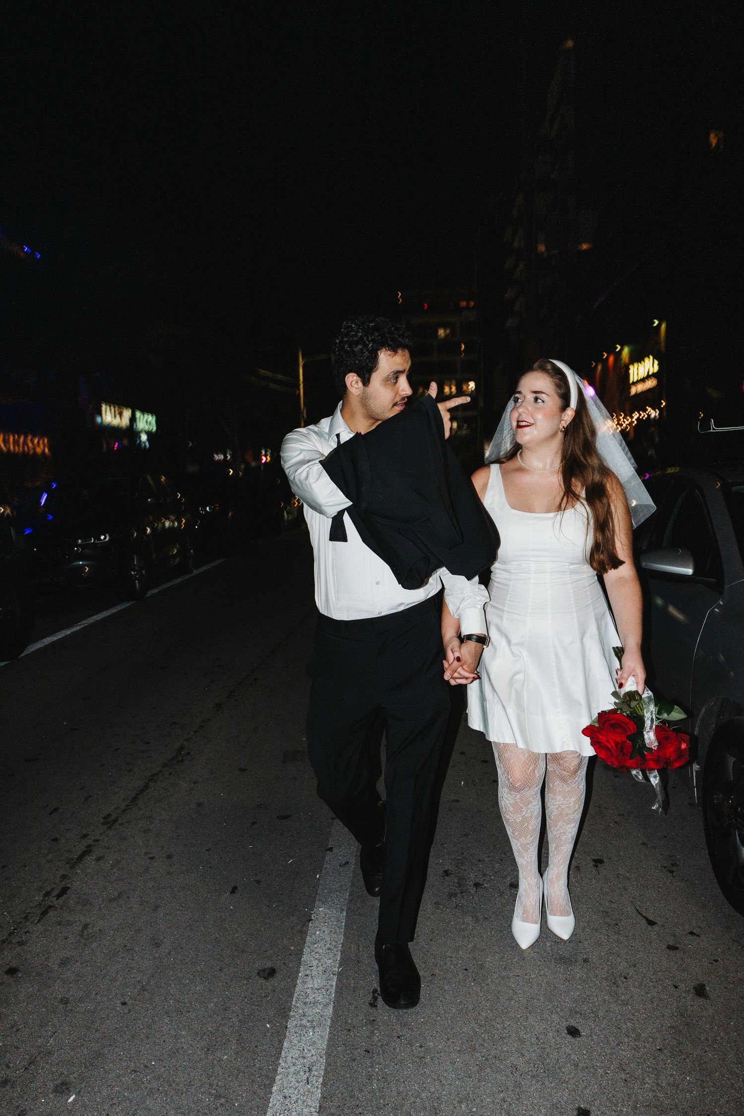 A couple in formal attire, with one person in a black suit and another in white, walks together on a dark street.