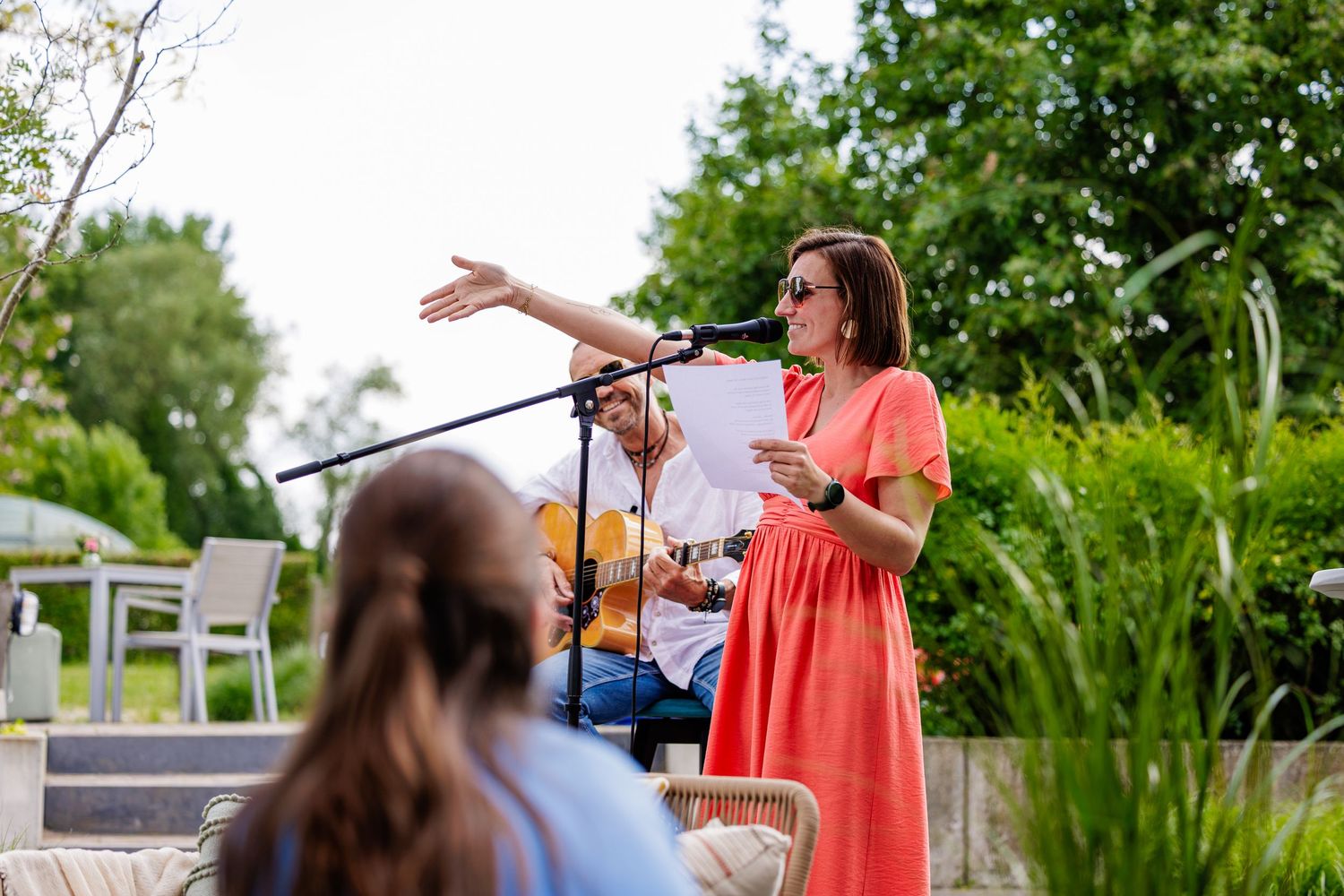 Musicians perform at an outdoor event on a sunny day with lush greenery in the background.