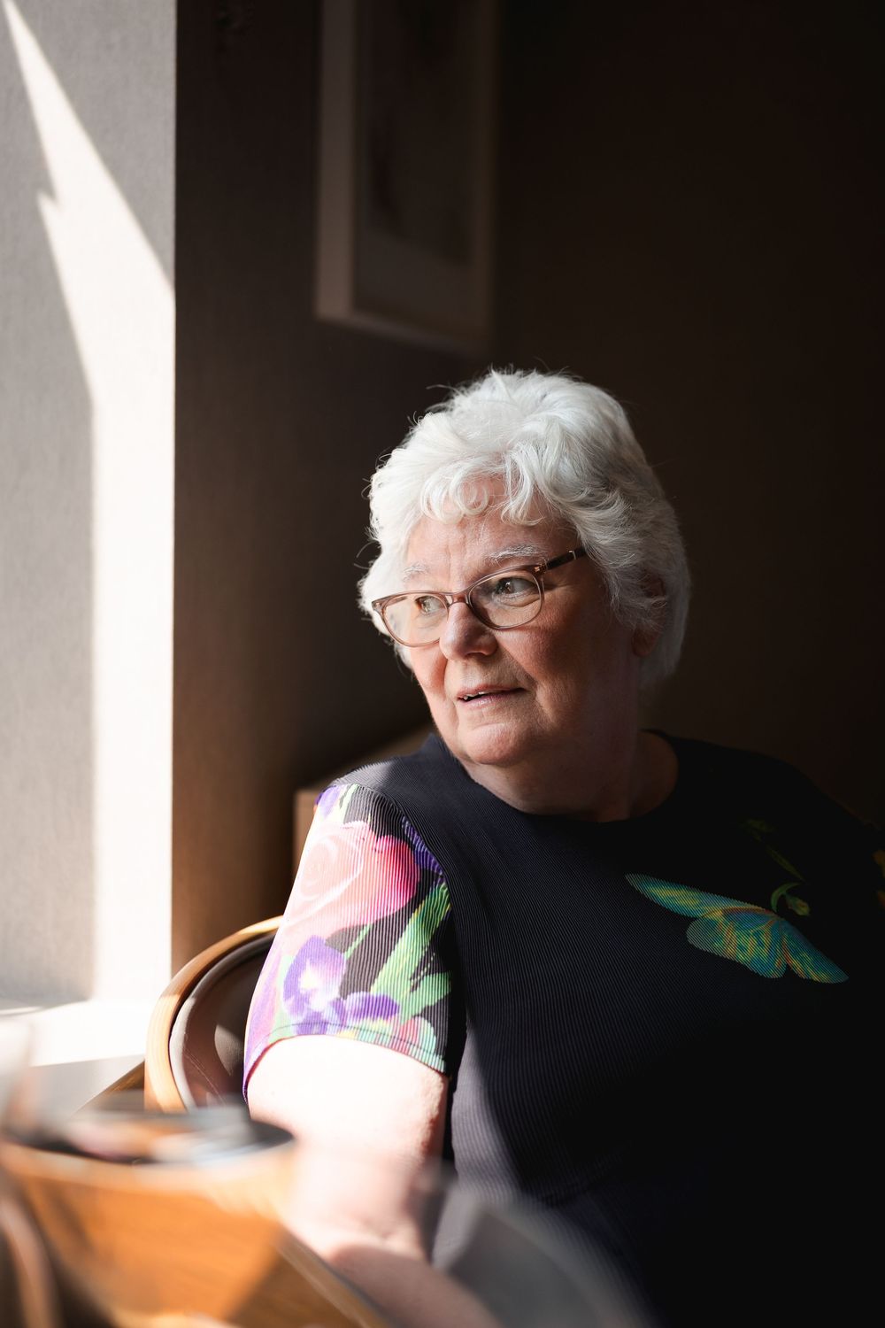 An elderly woman in glasses sits near a window with soft natural light creating a gentle shadowed portrait.