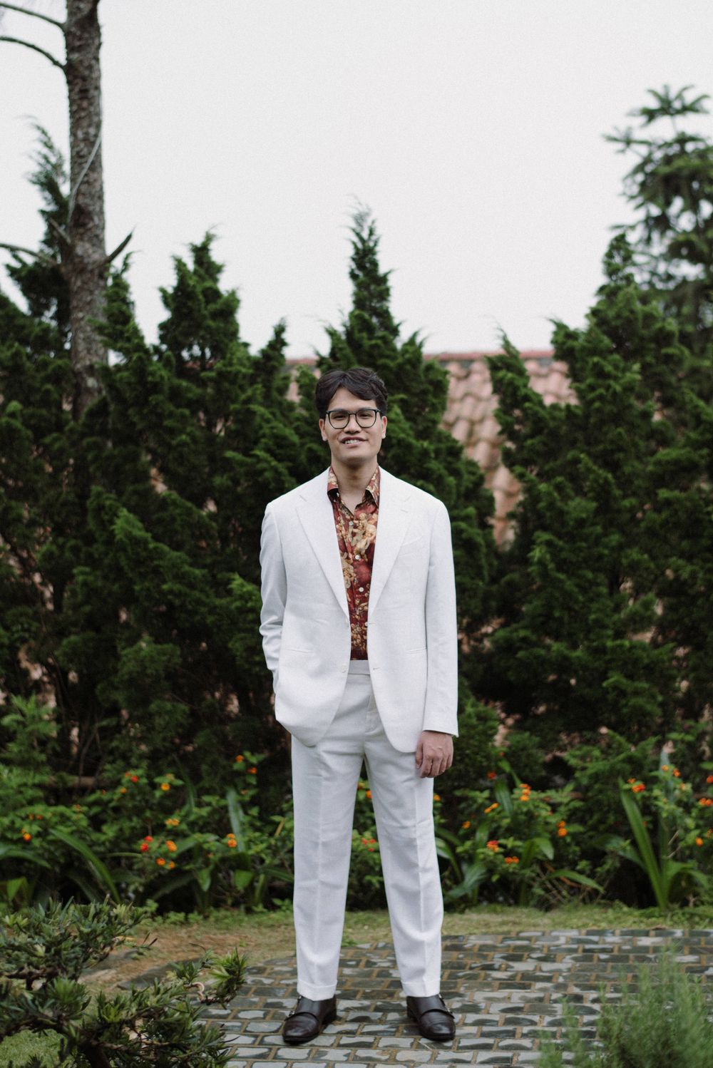 A groom in a crisp white suit poses against a backdrop of green cypress trees.