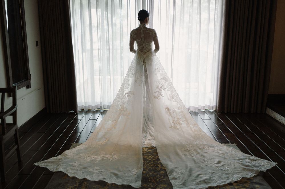 Silhouette of a wedding dress with flowing train against sheer curtains in a room.