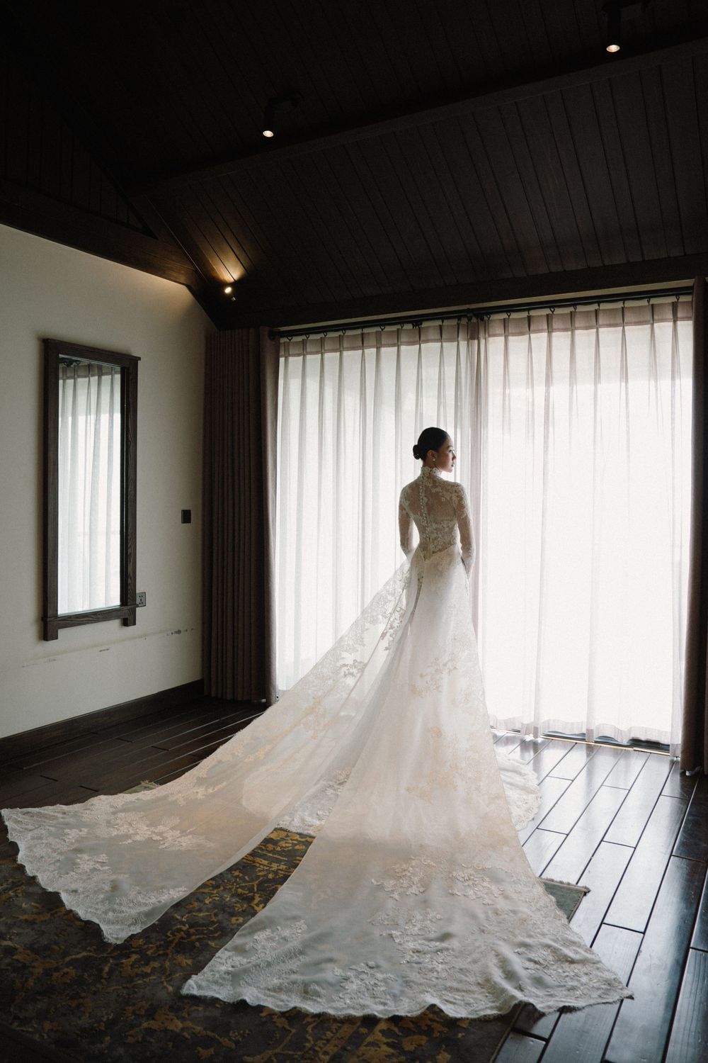 Long white wedding dress with lace train spread across dark wooden floor near window.