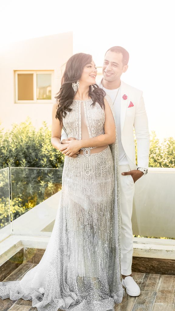 A couple in white formal wear embrace and laugh together on an outdoor terrace.