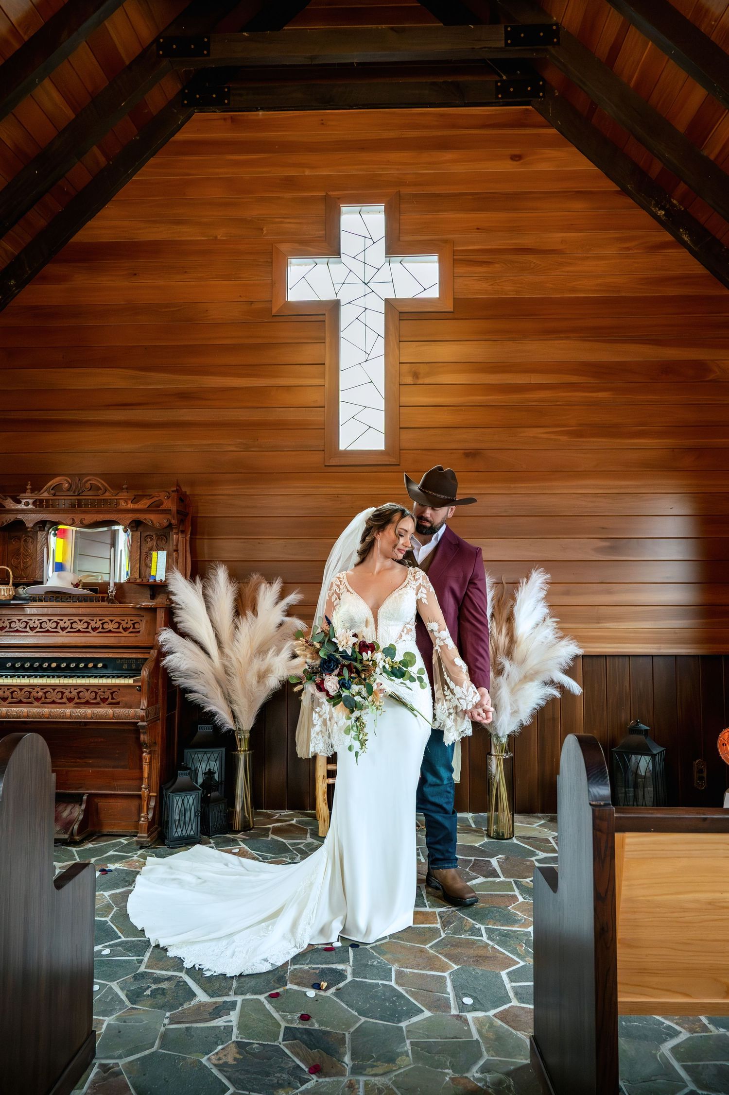 Bride and groom share a romantic moment in a rustic wooden chapel with a stained glass cross and pampas grass decor.