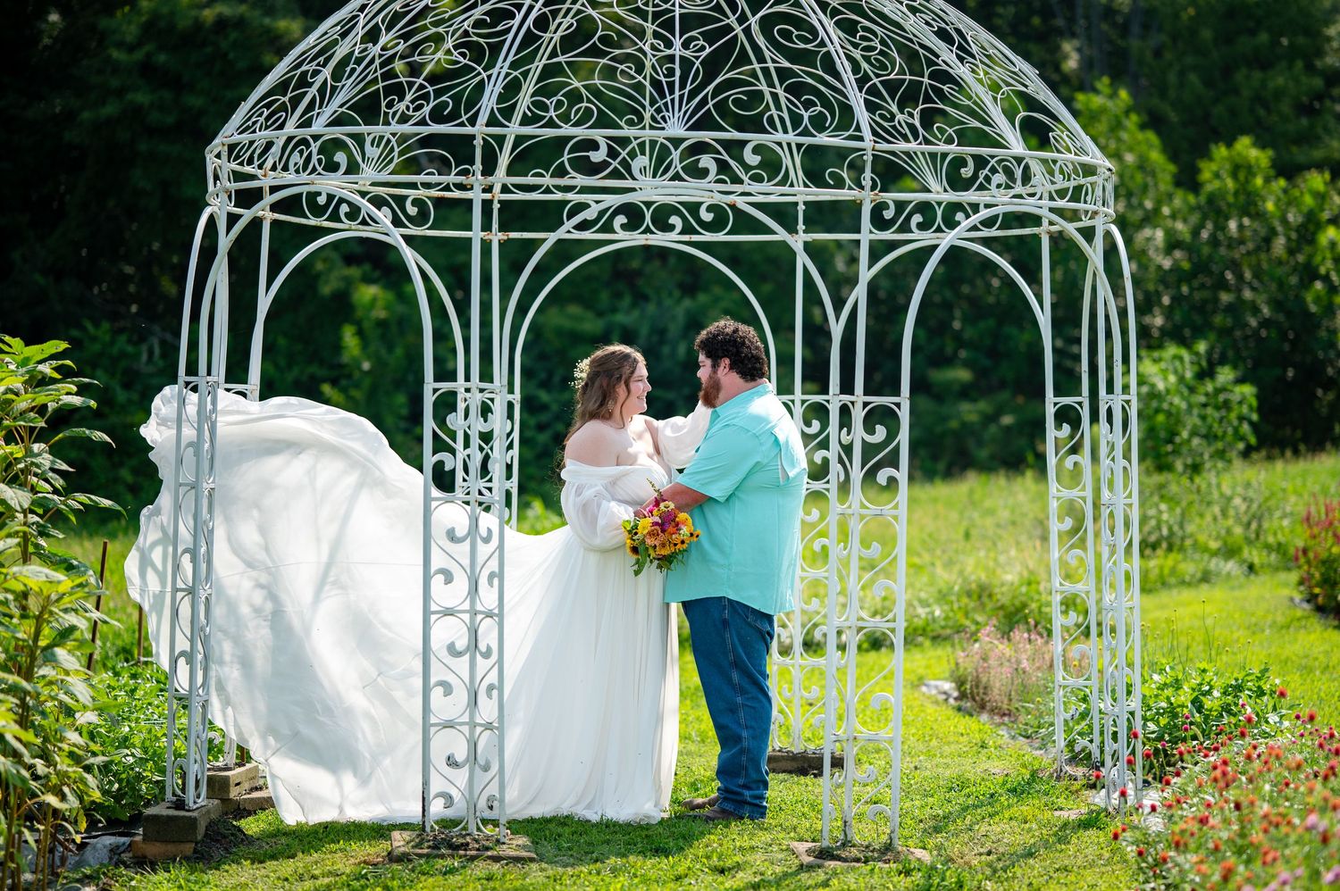 A couple stands together under an ornate white metal gazebo in a lush garden setting during a wedding ceremony.