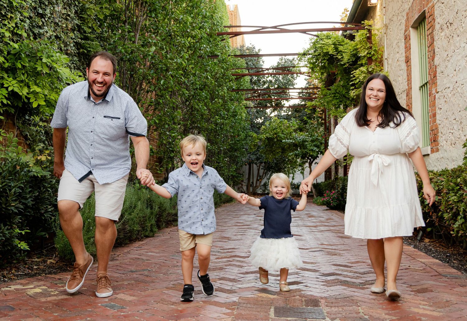 A happy family walks together on a brick path surrounded by lush greenery and gardens.