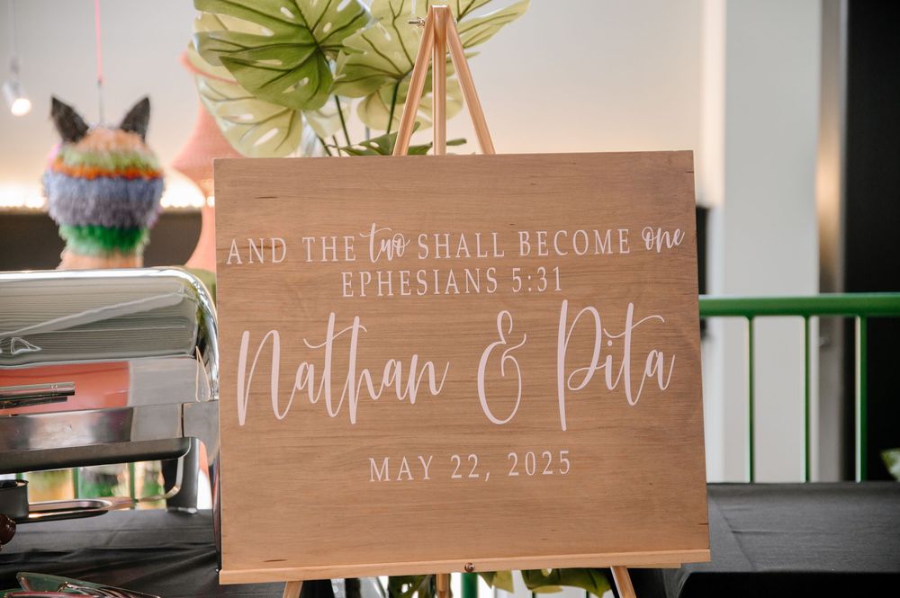 Wood wedding sign displays Ephesians 5:31 Bible verse and wedding details in elegant calligraphy at rustic ceremony.