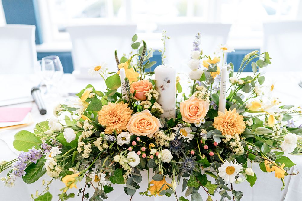 A lush floral arrangement with peach roses and white blooms cascades across a wedding reception table setting.