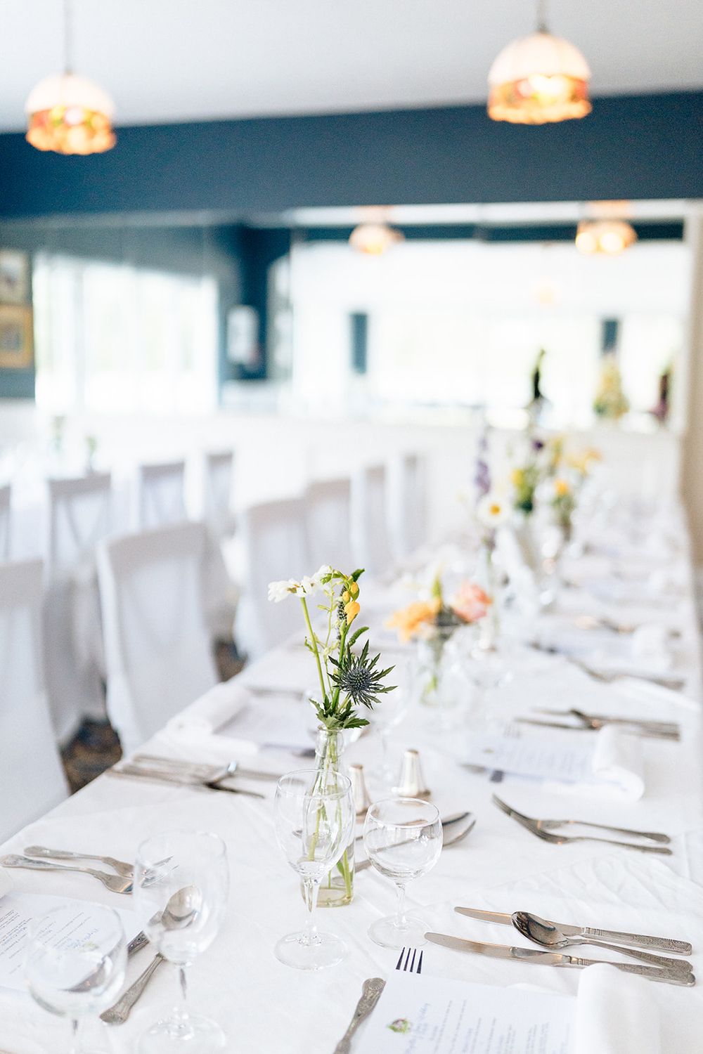 Elegant minimalist wedding reception table setting with white linens, delicate flower vases and warm pendant lighting above.