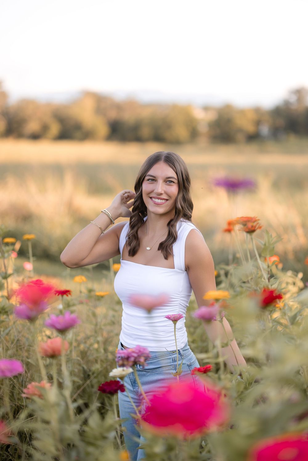 Woman in white tank top and jeans stands among colorful wildflowers in a golden field during sunset.