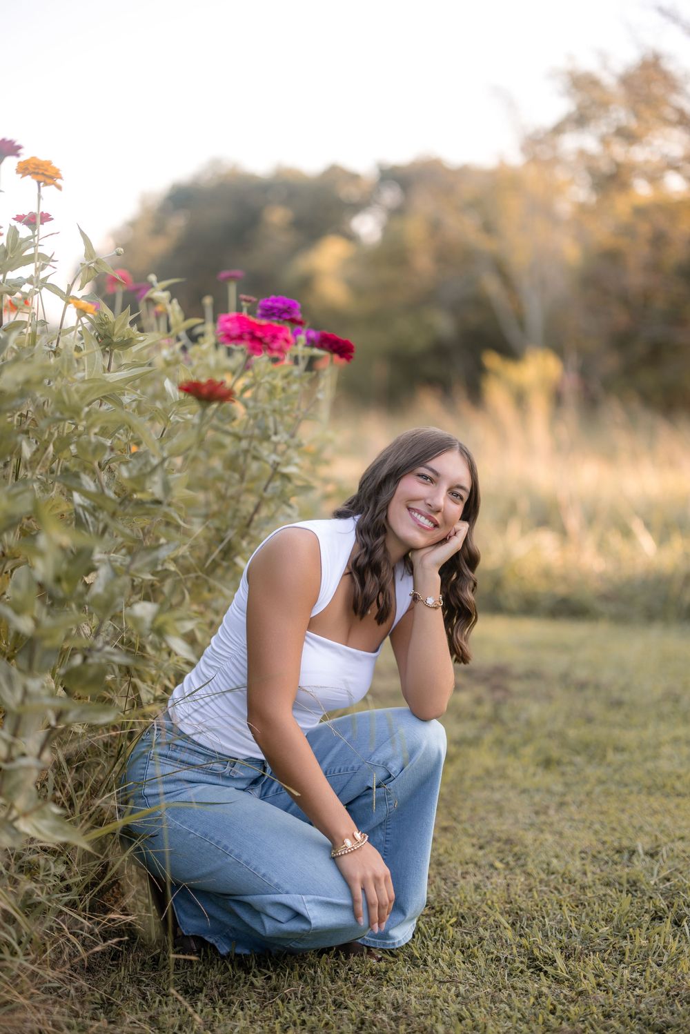 A stylish woman in jeans and white top poses near blooming pink flowers in a golden-hour outdoor setting.