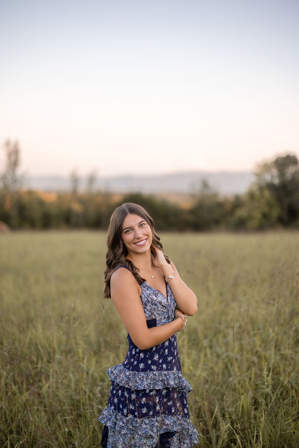 A person in a blue floral dress sits in a sunny field at sunset while smiling and touching their hair.
