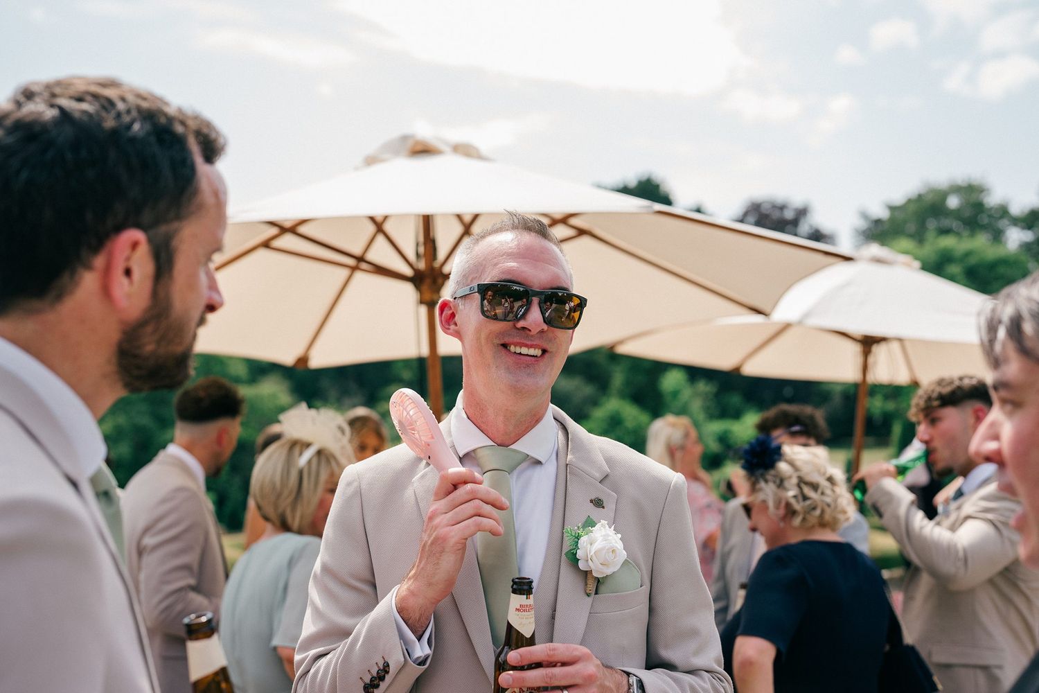 A group of guests enjoying themselves at an elegant outdoor wedding reception under beige umbrellas on a sunny day.