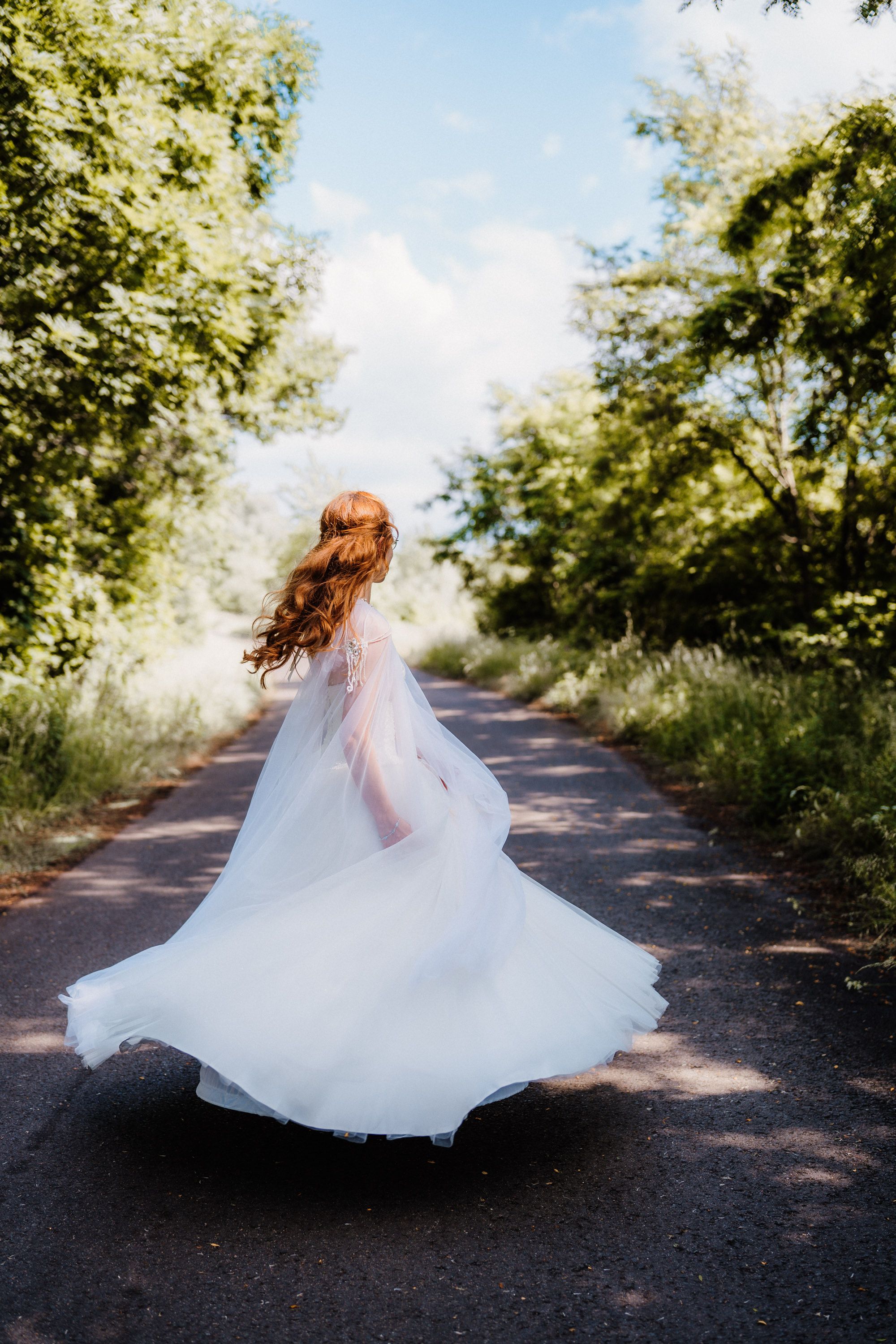 Bride twirling in flowing white dress on a tree-lined path on her wedding day.