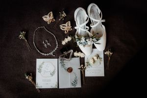 Wedding details flatlay with white shoes, invitation cards, jewelry and butterfly decorations on dark background.
