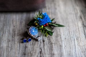 Wedding accessories featuring blue flowers and watch arranged on wooden surface.